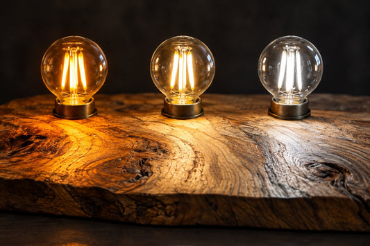 Três lâmpadas LED de filamento acesas sobre uma tábua de oliveira, em luz âmbar, branco quente e branco frio, mostrando como cada cor de luz muda o veio da madeira
